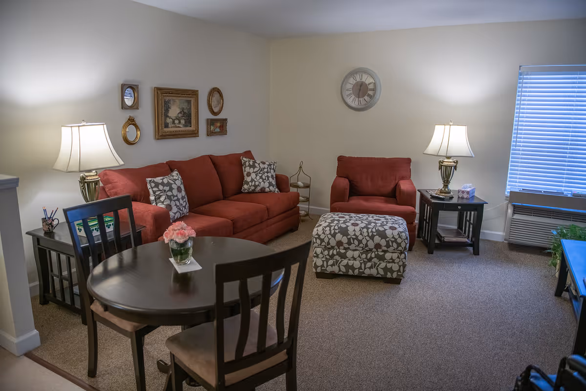 Cozy living room with a red sofa and armchair, patterned ottoman, small dining table, lamps, wall art, and a window with blinds.
