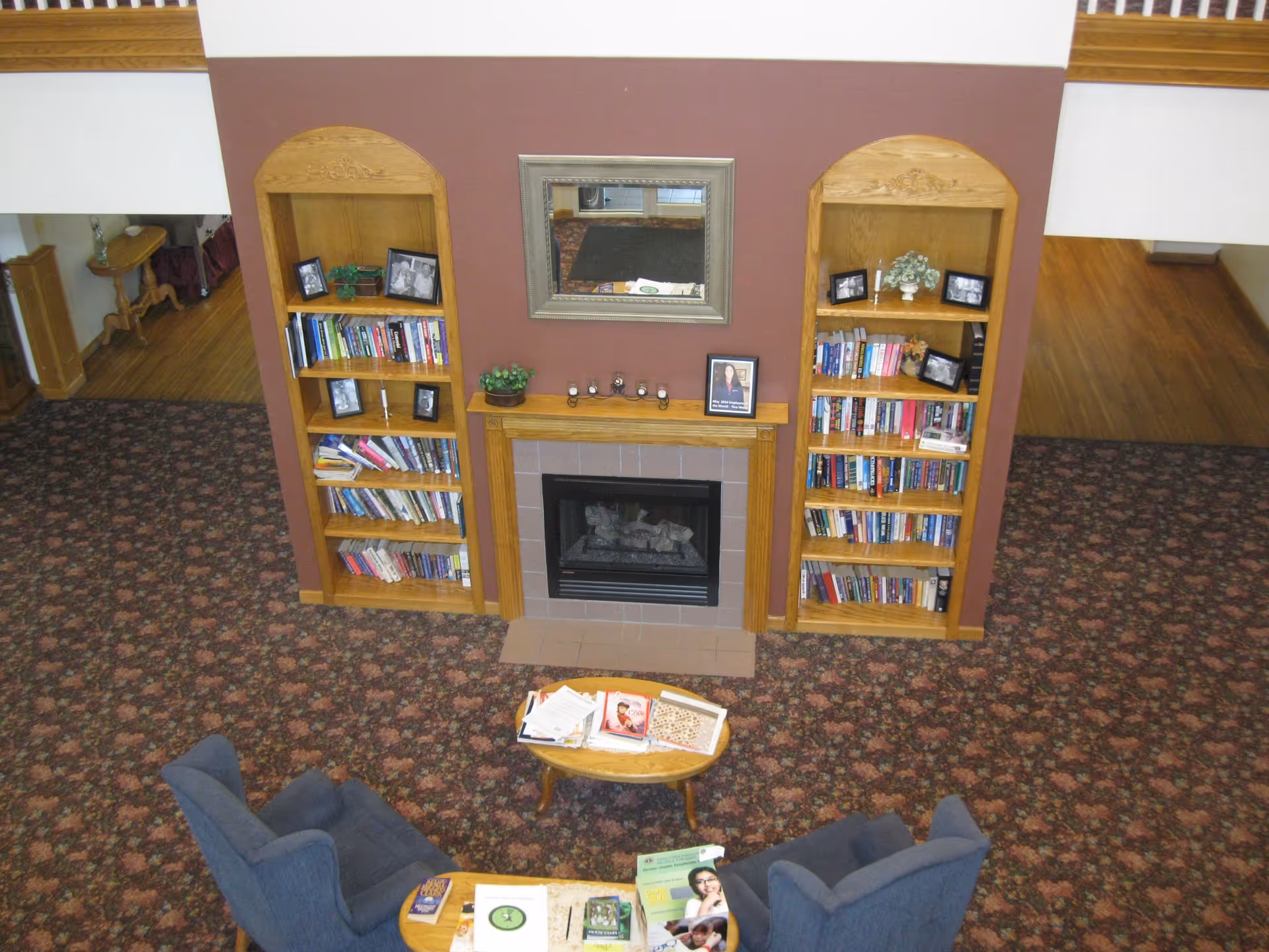 An interior lounge with a central fireplace flanked by wooden bookshelves, two blue armchairs and a coffee table on patterned carpet.
