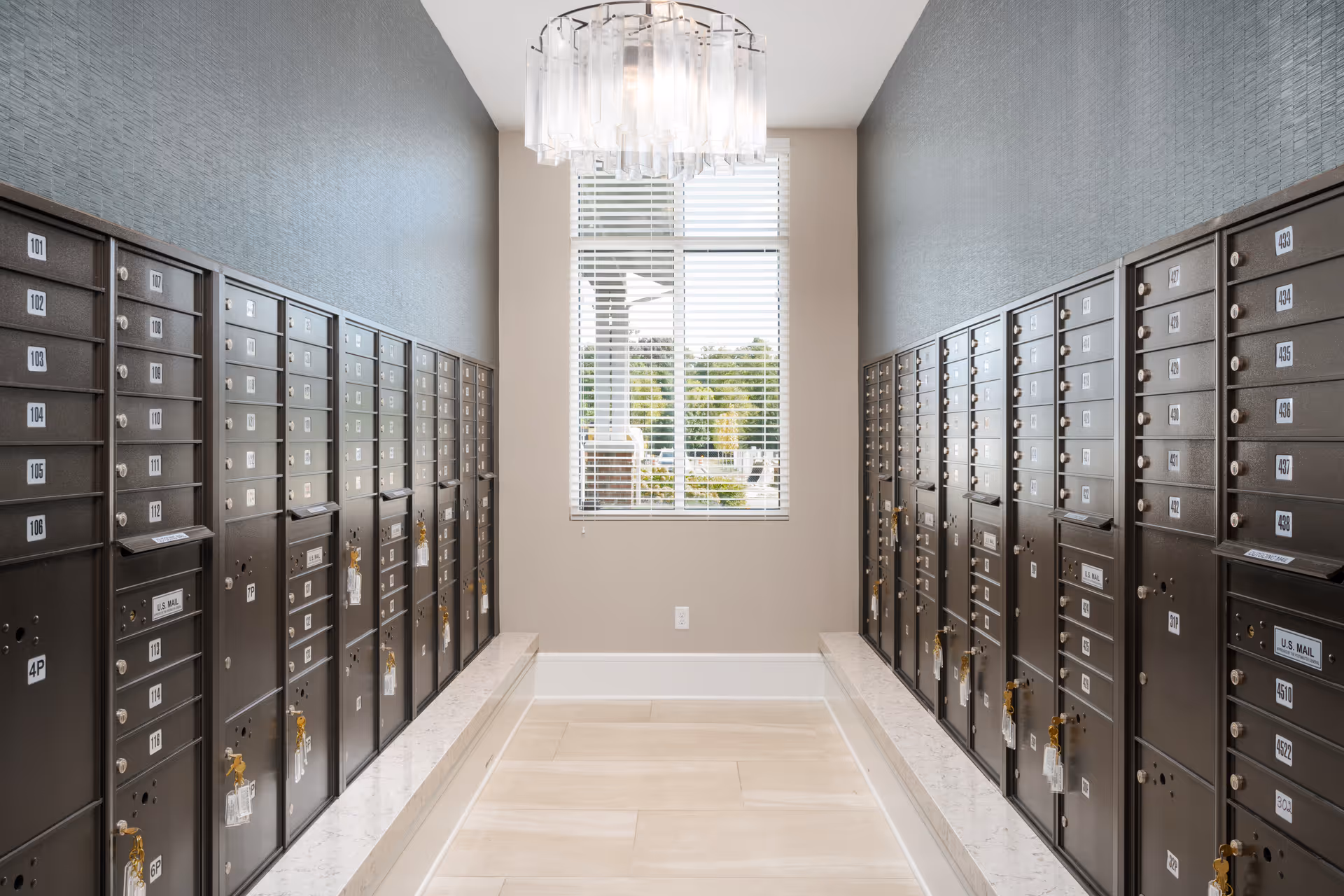 Interior mailroom with rows of numbered mailboxes along both walls, a window at the end, and a chandelier overhead.