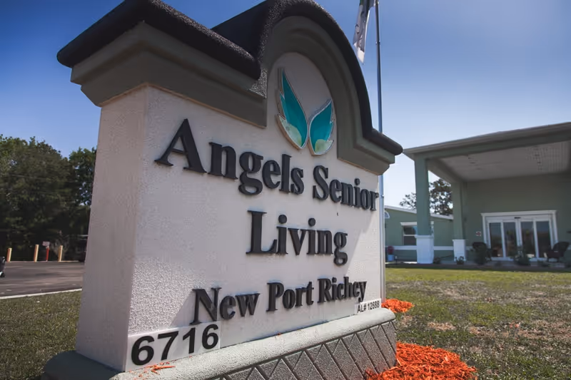 Outdoor view of a large sign for Angels Senior Living New Port Richey with the building entrance and lawn visible in the background under a clear blue sky.