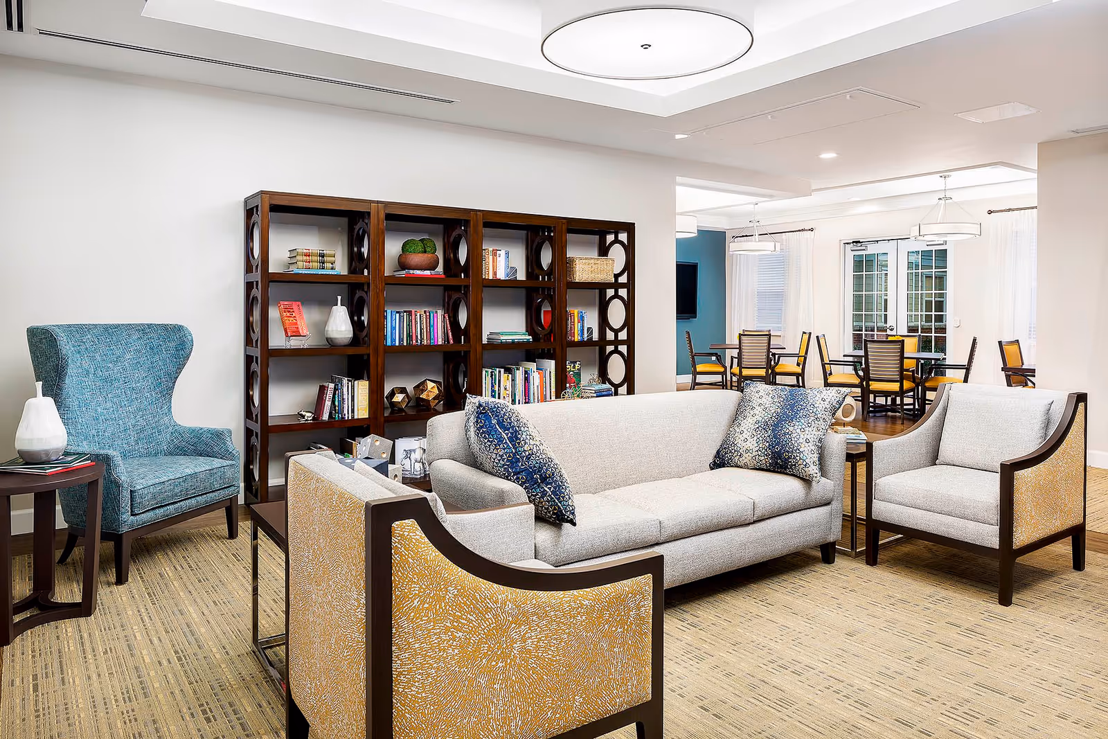 A bright and spacious living room area in a senior living facility featuring a light gray sofa with blue patterned pillows, two armchairs with yellow patterned upholstery, a blue wingback chair, a dark wooden bookshelf filled with books and decorative items, and a dining area with tables and chairs in the background near large windows with white curtains.
