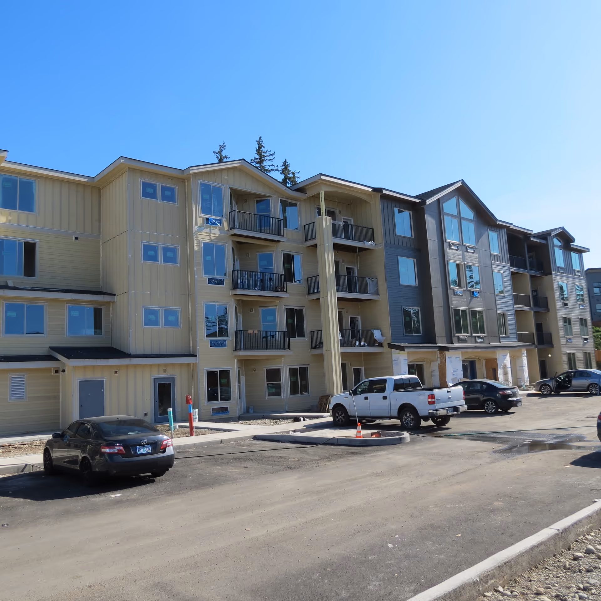 Four-story modern apartment building with balconies and parked cars in front under a clear blue sky.