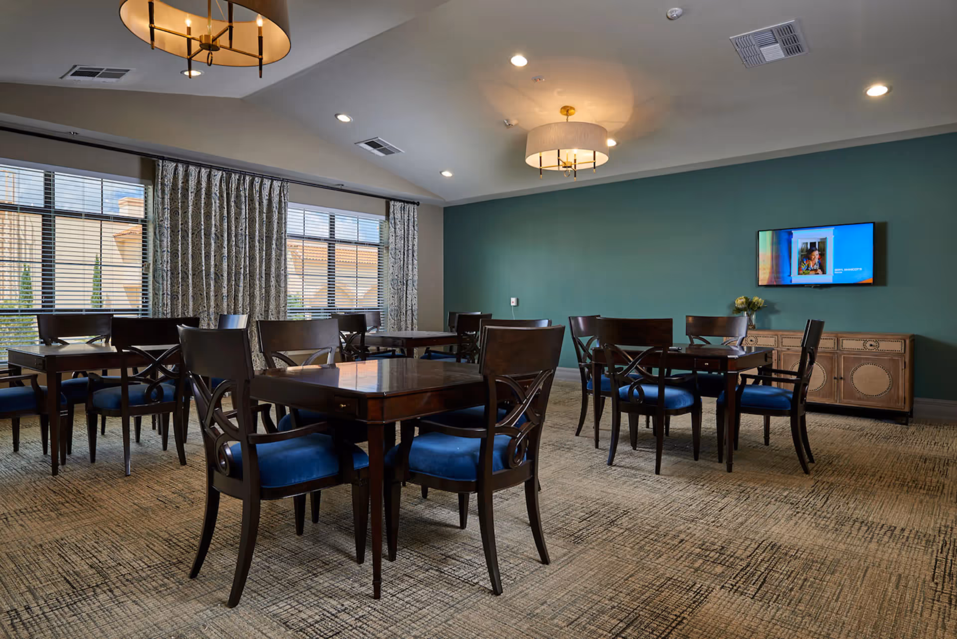 A dining room with several dark wooden tables and chairs with blue cushions. The room has a green accent wall with a mounted flat-screen TV and a wooden sideboard beneath it. Large windows with patterned curtains allow natural light to enter. The ceiling features recessed lighting and two modern chandeliers.