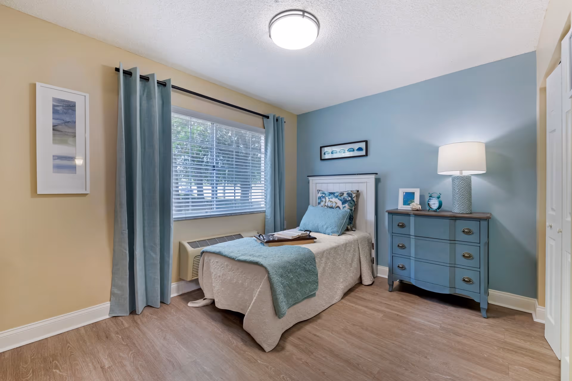 A neatly arranged bedroom with a single bed covered in a white quilt and light blue throw blanket and pillows. There is a blue dresser with a lamp, a small framed picture, and a clock on top. The walls are painted light blue and beige, with a window covered by light blue curtains and white blinds. The floor is wooden.