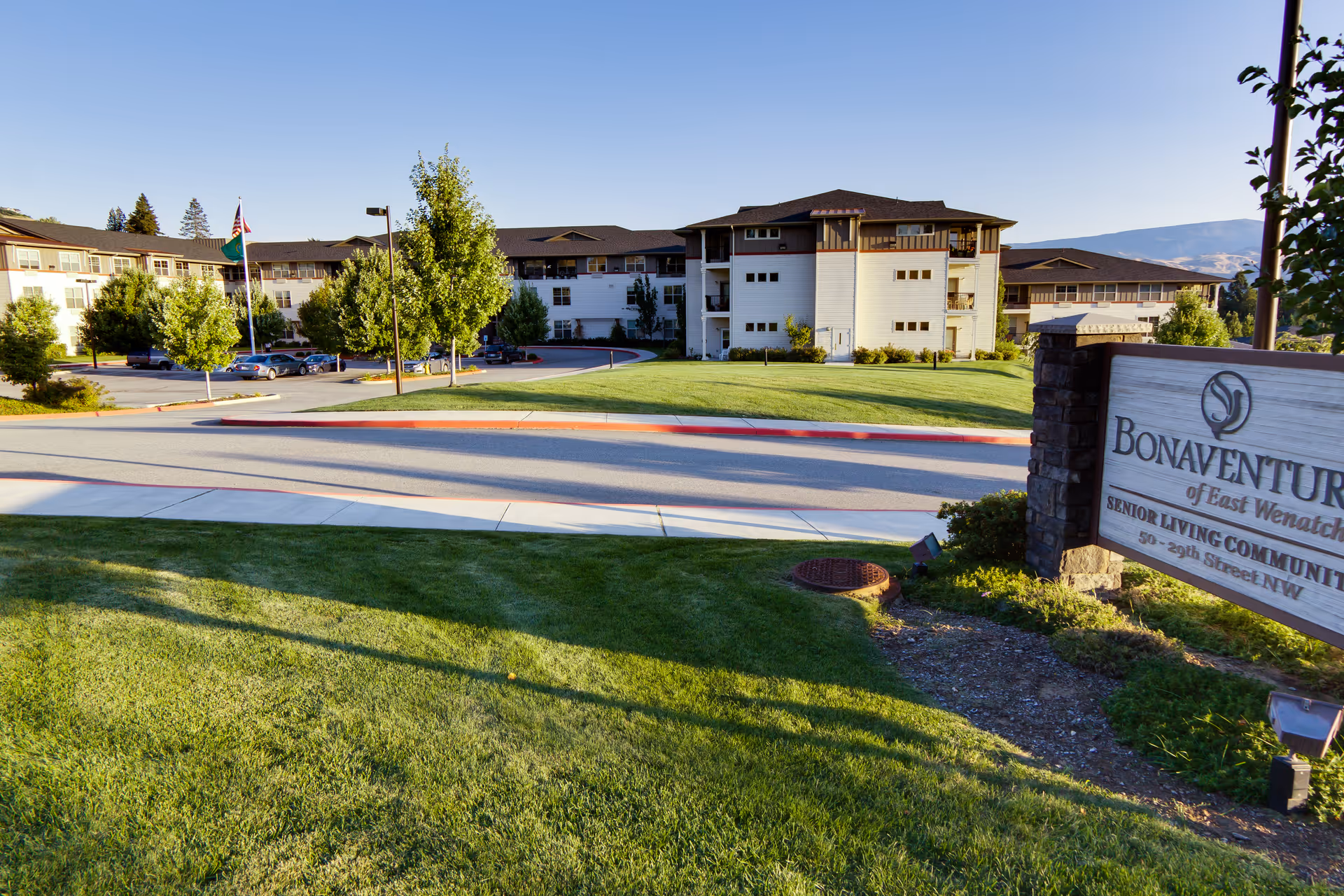 Exterior view of Bonaventure of East Wenatchee senior living community building with a well-maintained lawn, trees, a parking area with cars, and a sign displaying the facility's name and address.