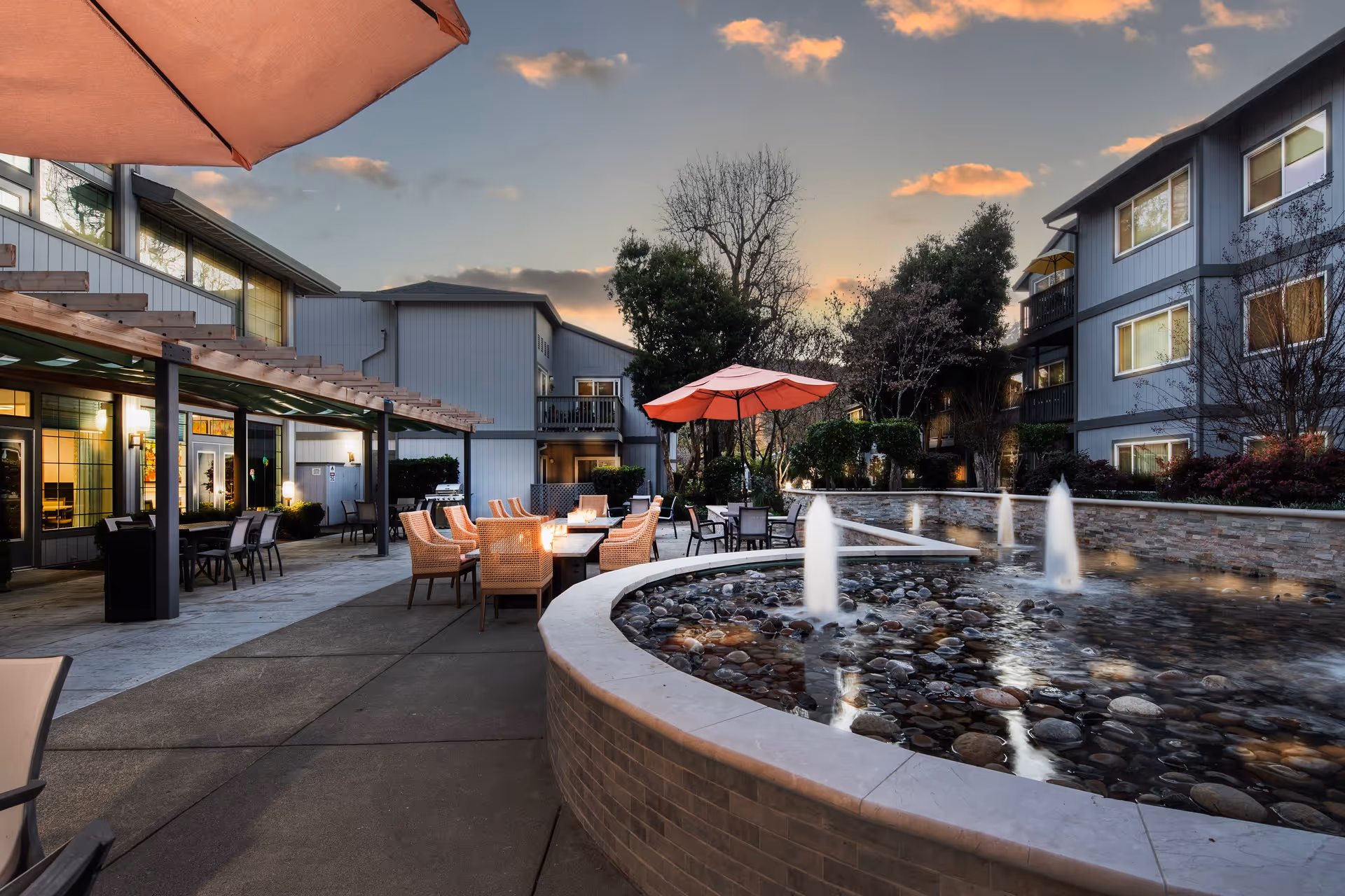 Outdoor patio area at sunset with seating arrangements including chairs and tables under umbrellas, a water feature with fountains, and surrounding buildings with windows and balconies.