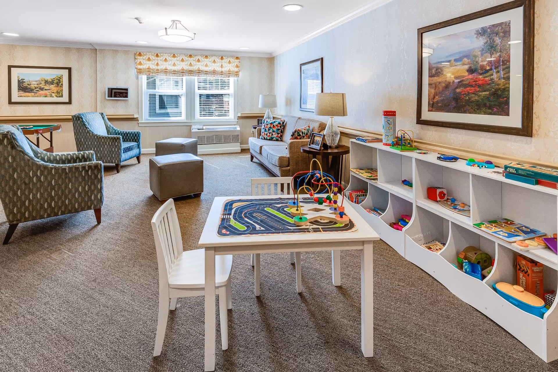 A bright and cozy common area in a senior living facility featuring a small white table with a bead maze toy and a race track play mat, a white chair, a long white shelf filled with various toys and games, two patterned armchairs, two ottomans, a beige sofa with colorful pillows, a side table with a lamp, framed landscape paintings on the walls, and a window with patterned blinds letting in natural light.