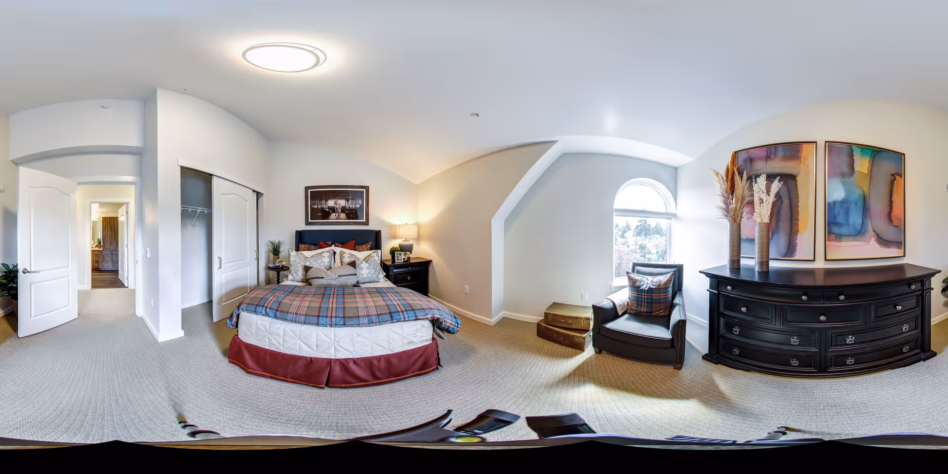 A bright and spacious bedroom with a bed dressed in a plaid blanket and multiple pillows. To the right of the bed is a black nightstand with a lamp and decorative items. A comfortable armchair with a plaid pillow sits near a window with an arched top, letting in natural light. Next to the armchair is a large black dresser with two colorful abstract paintings hanging above it. The room has light-colored walls and carpet, with an open door leading to a hallway and closet with sliding doors.