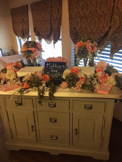 A decorated buffet table indoors with floral arrangements and a small chalkboard sign reading "Mother's Day sweets" in front of window draperies.
