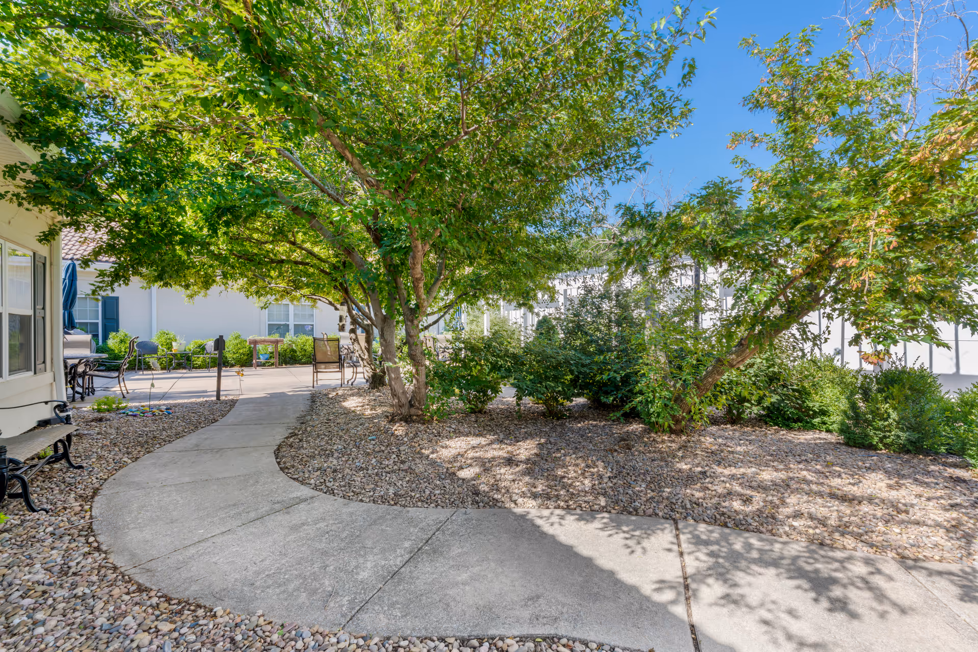 Sunlit courtyard walkway with trees, gravel beds, and patio seating beside a building.