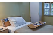 A single bed in a simple, clean room with white pillows and beige bedding. A wooden overbed table with magazines rests on the bed. There is a window with multiple panes letting in natural light, and a closed door is visible in the background.