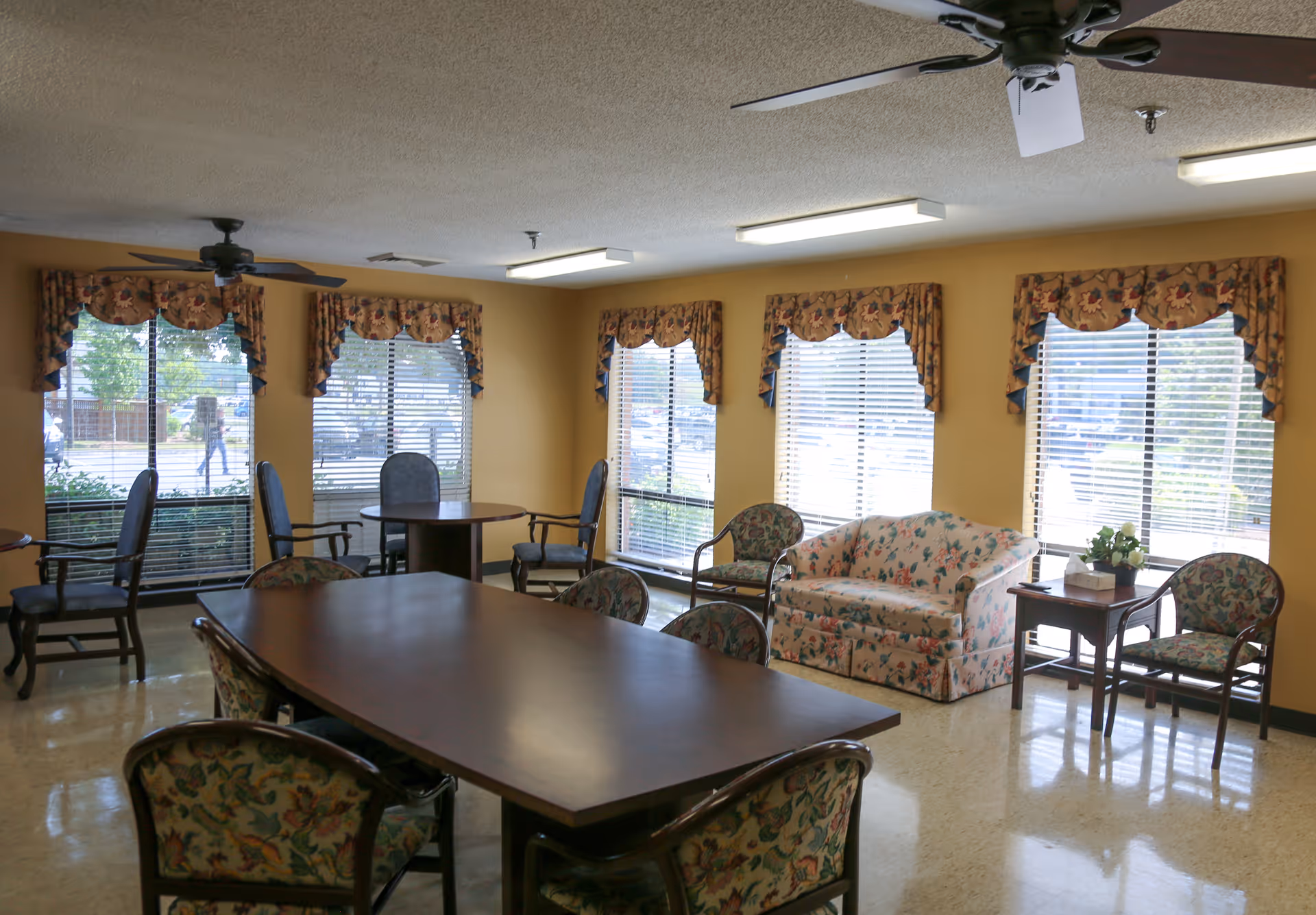 A well-lit room with large windows covered by floral valances and blinds. The room contains a long wooden table surrounded by floral upholstered chairs, a floral upholstered loveseat, additional chairs, and small side tables. Ceiling fans and fluorescent lights are mounted on the ceiling.