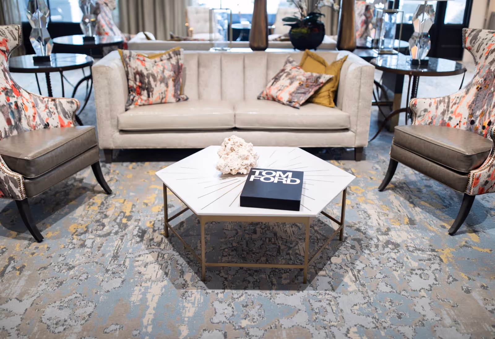 A modern living room area with a beige sofa adorned with colorful patterned pillows, two matching armchairs with a similar pattern, and a hexagonal coffee table with a decorative coral piece and a Tom Ford book on top. The floor is covered with a patterned carpet in shades of blue, gray, and yellow.