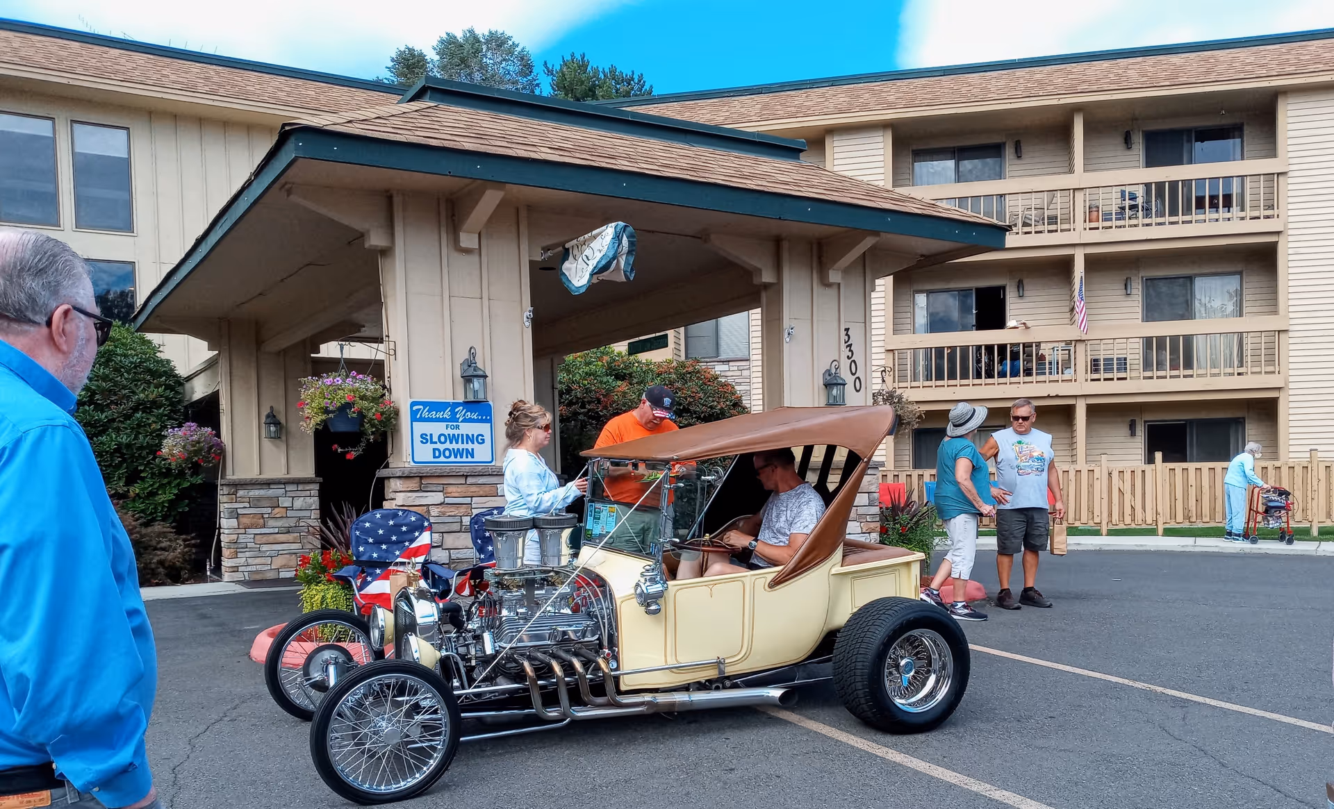 A group of people gathered outside the entrance of Ponderosa Assisted Living Community. A vintage cream-colored car with a brown roof is parked in front. The building has multiple balconies and a sign near the entrance reads 'Thank You for Slowing Down.' Some people are standing and conversing, while an elderly person with a walker is visible in the background.