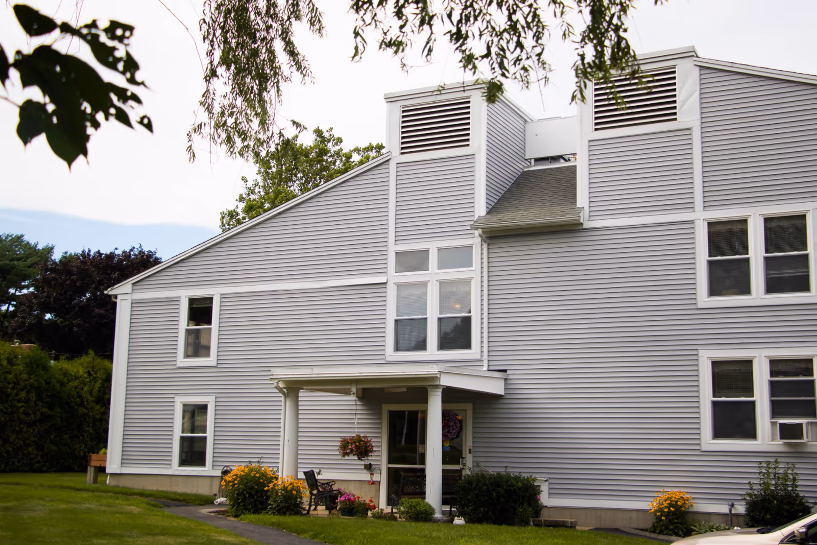 Gray two-story building with white trim, a small covered entrance, and landscaping with flowers and lawn.