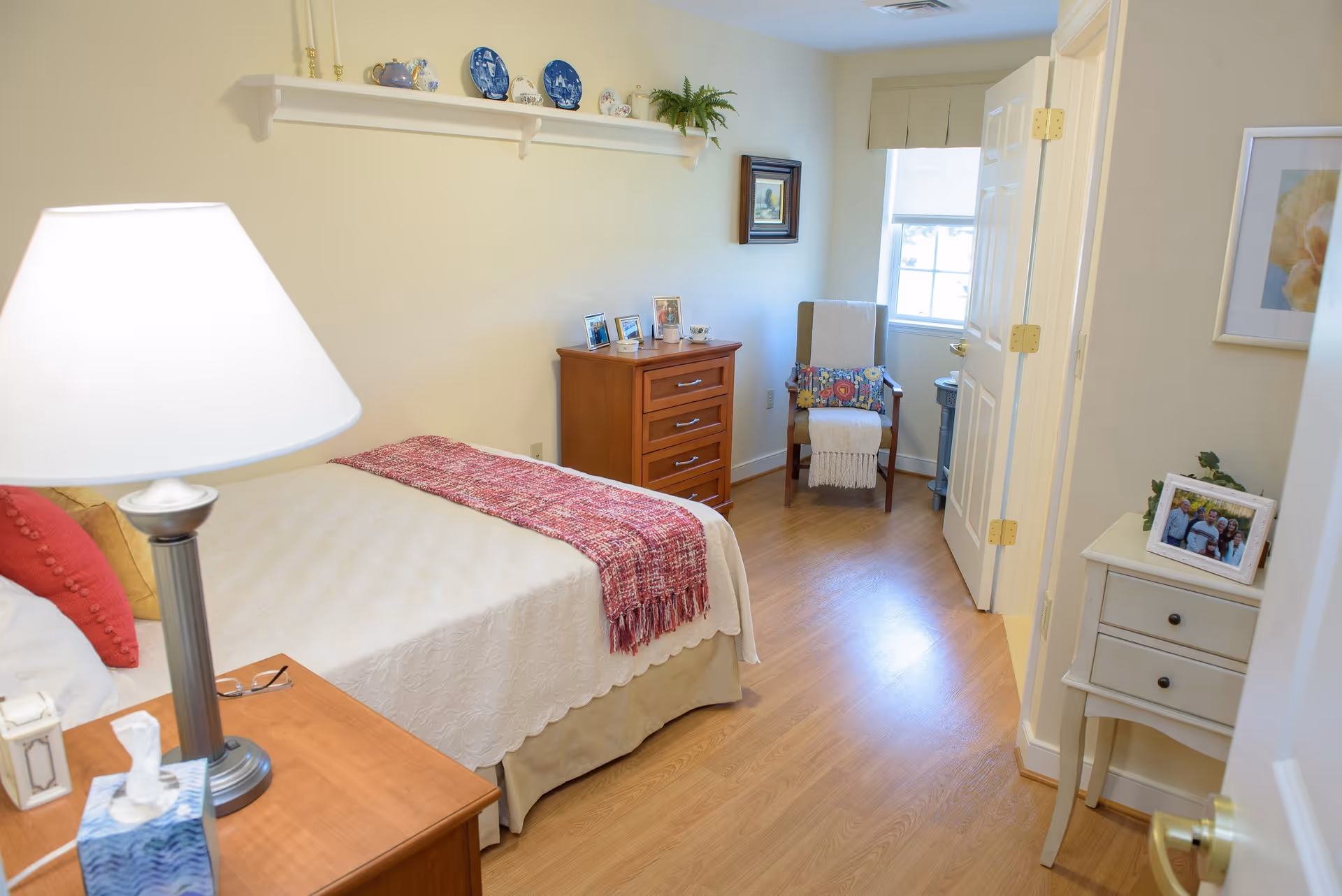 A cozy bedroom with a single bed covered in a white quilt and a red throw blanket. There is a wooden nightstand with a lamp and tissue box next to the bed. A wooden dresser with framed photos is against the wall, and a chair with a colorful cushion and a white throw is placed near a window. The room has light-colored walls, wooden flooring, and decorative plates on a shelf above the dresser.