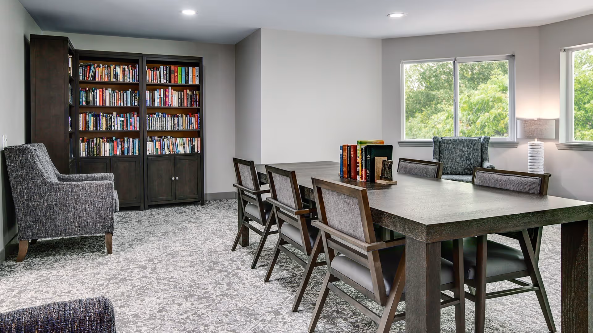 A quiet reading or meeting room with a large wooden table surrounded by six chairs. There are two armchairs near the windows, which let in natural light and show green trees outside. A tall bookshelf filled with books is against the wall, and a table lamp is placed near one of the windows.