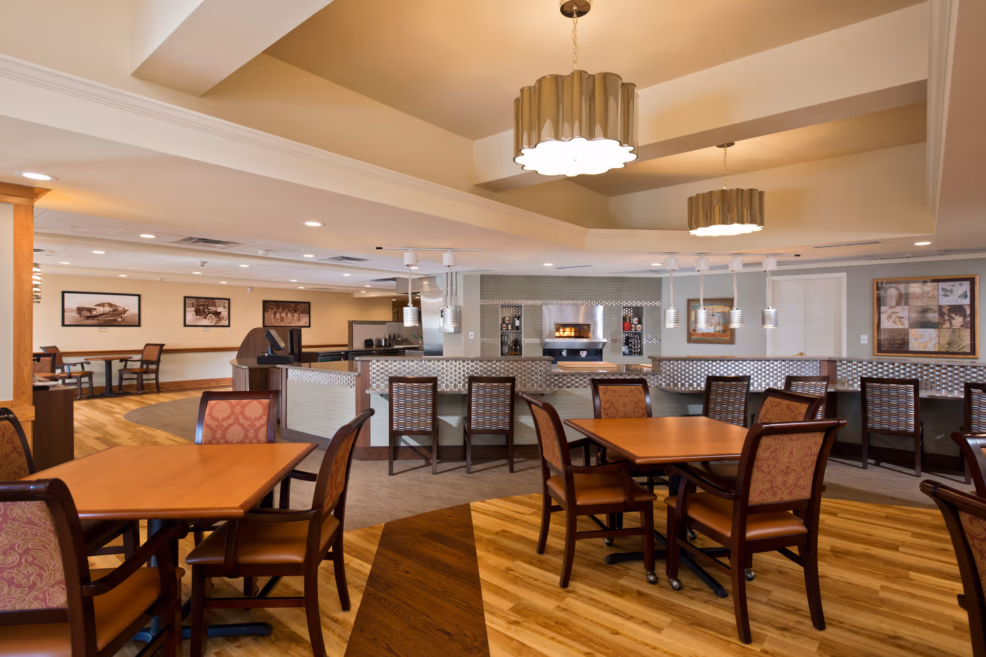 A spacious dining area in a senior living facility with wooden tables and cushioned chairs arranged neatly. The room features a modern ceiling with recessed lighting and two large pendant lights. In the background, there is a counter with bar stools and a built-in oven or fireplace, along with framed artwork on the walls.