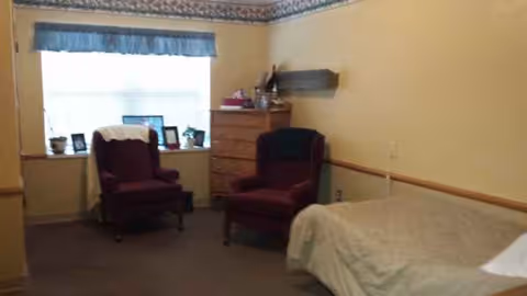 Bedroom in a senior living facility with two burgundy chairs, a dresser, and a bed by window.