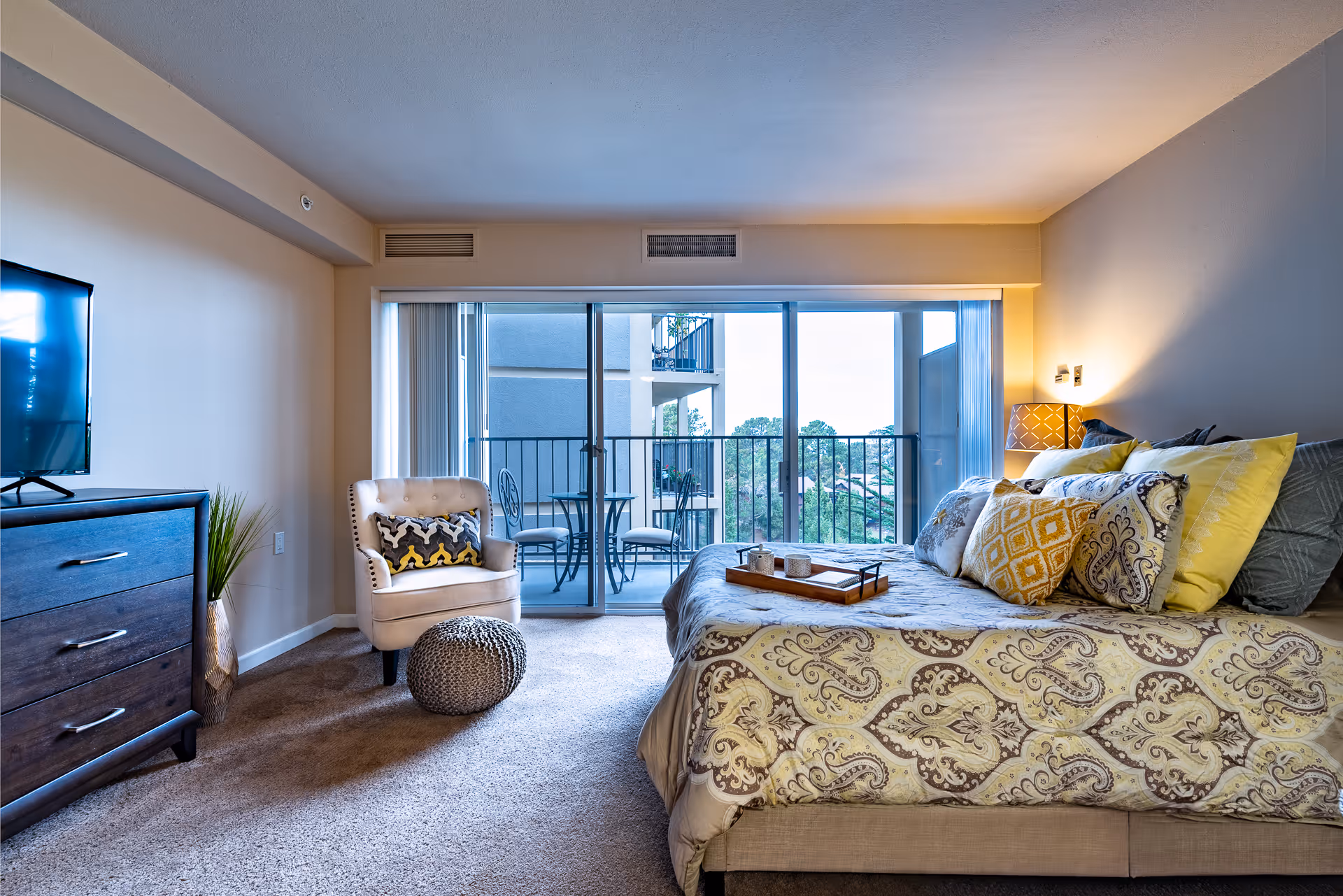 Well-lit bedroom with a decorated bed, armchair and dresser, and sliding glass doors opening to a balcony with outdoor seating.