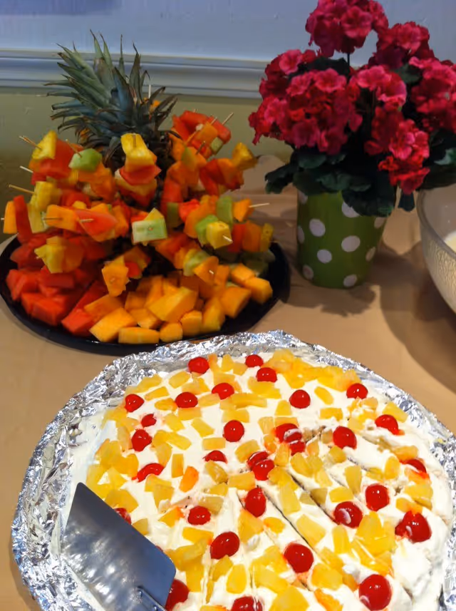 A table with a fruit pizza topped with whipped cream, cherries, and pineapple chunks, next to a platter of colorful fruit skewers arranged around a pineapple base, and a green polka dot vase with bright pink flowers.