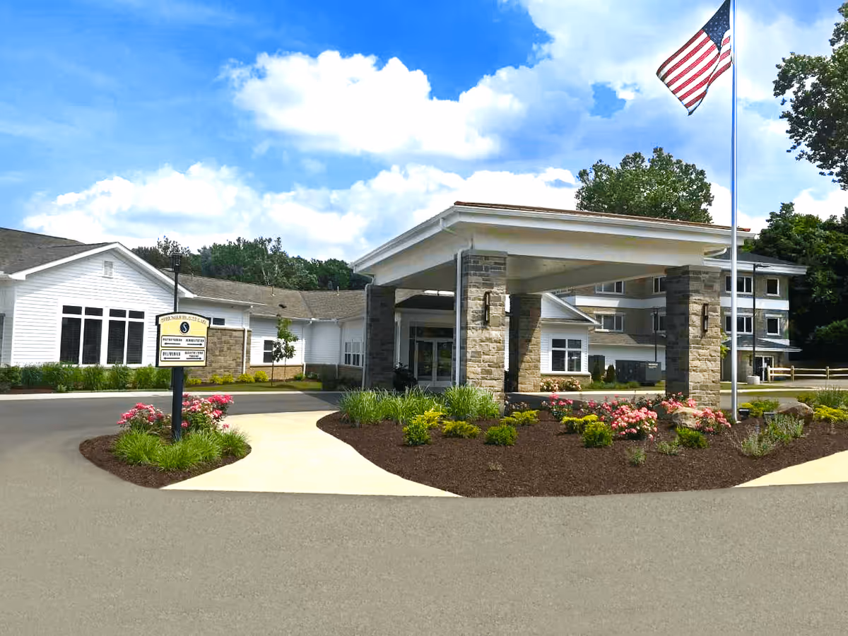 Exterior view of Sprenger Health Care Amherst Manor Retirement Community showing the entrance with a covered drop-off area supported by stone pillars, landscaped flower beds with pink flowers and green shrubs, an American flag on a tall flagpole, and a clear blue sky with some clouds.