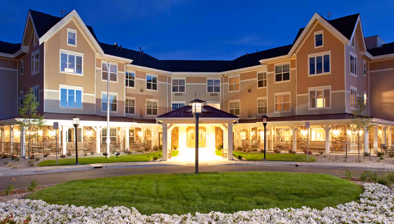 Exterior view of MorningStar Assisted Living & Memory Care at Jordan building at dusk, showing a three-story structure with multiple windows, warm lighting, a covered entrance, green lawn, and white flowers in the foreground.