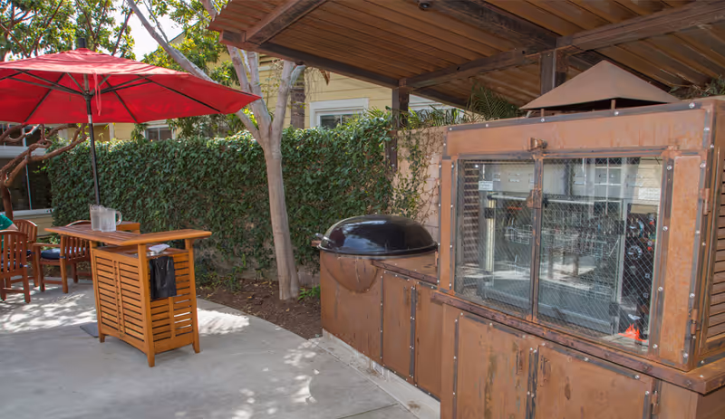 Outdoor patio area with a wooden serving cart under a red umbrella, surrounded by wooden chairs. There is a large rust-colored outdoor grill or smoker with a glass door on the right side, and a green ivy-covered wall in the background.