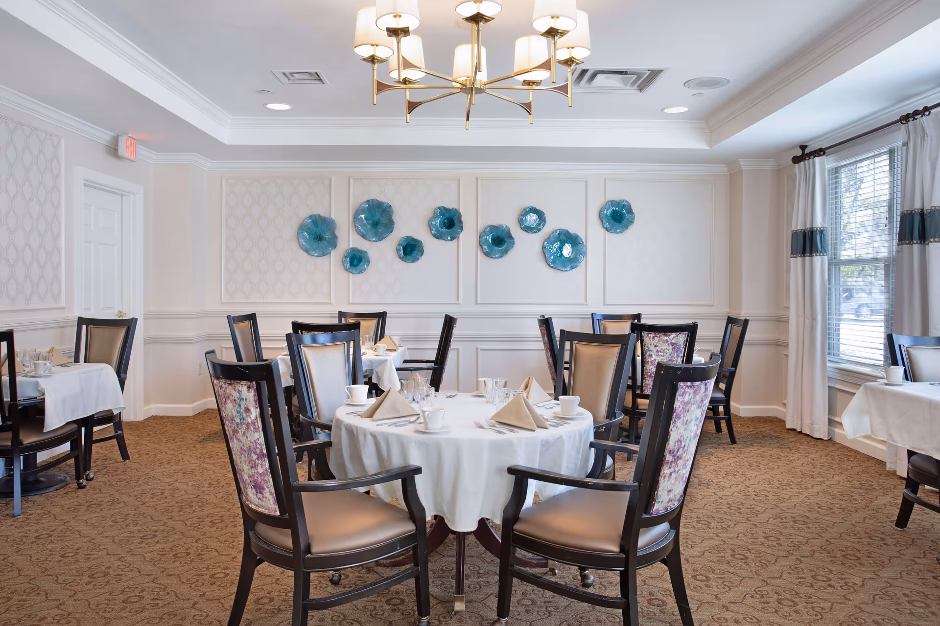 A bright and elegant dining room with round tables covered in white tablecloths, set with cups, glasses, and folded napkins. The chairs have dark wooden frames with beige and floral upholstery. The walls are decorated with blue ceramic wall art, and large windows with white curtains allow natural light to fill the room. A modern chandelier hangs from the ceiling.