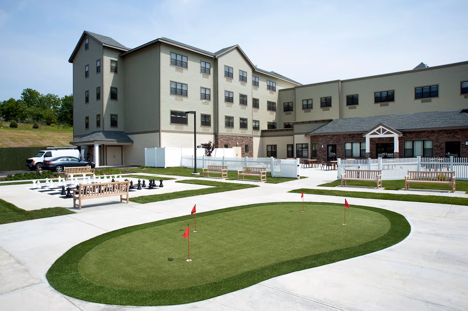Outdoor area of Braemar at Wallkill senior living facility featuring a putting green with four red flags, several wooden benches, a large outdoor chess set, and a multi-story building in the background under a clear sky.