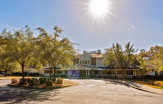 Front exterior of a senior living facility with a circular driveway, trees, and bright sun overhead.