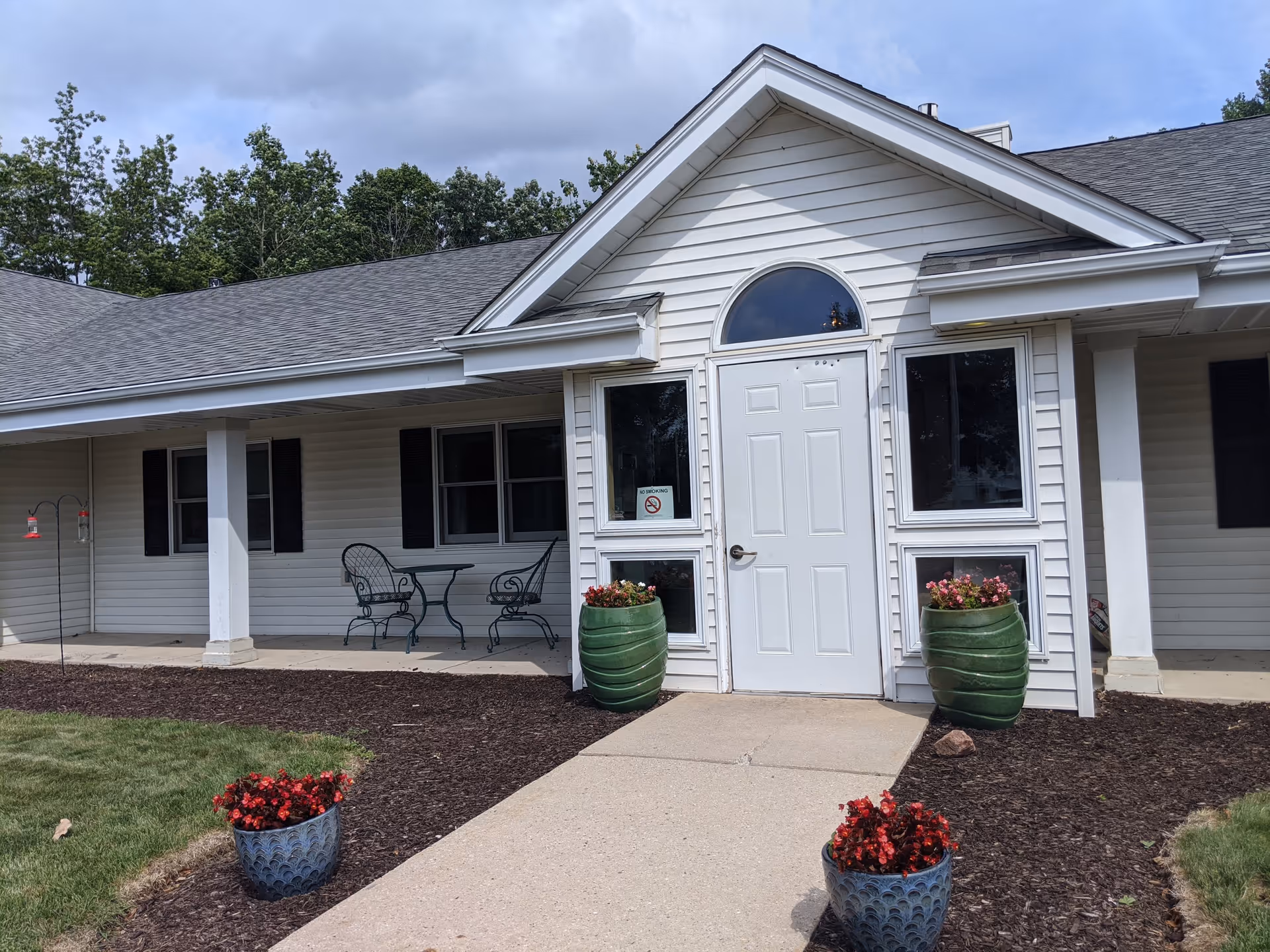 Exterior view of a single-story building with white siding and a gray shingled roof. The entrance features a white door with an arched window above it, flanked by two windows. There are two large green planters with flowers on either side of the door and two smaller blue planters with red flowers along the walkway. A small patio area to the left has a metal table and two chairs. Trees and a cloudy sky are visible in the background.
