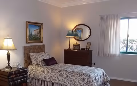A neatly furnished single bedroom with a twin bed, patterned bedspread, a dresser with lamp and oval mirror, framed artwork, and a curtained window.