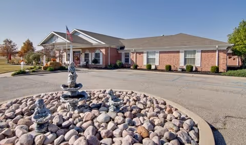 Single-story brick building with a gabled roof and an American flag near the entrance. In the foreground, there is a circular rock garden with three small tiered water fountains. The sky is clear and blue.