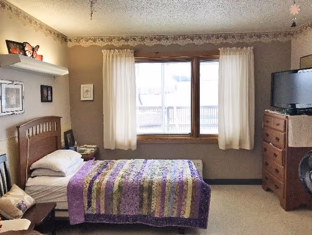 Furnished single bedroom with a bed covered by a purple quilt, window with white curtains, dresser with a TV, and a bedside table.