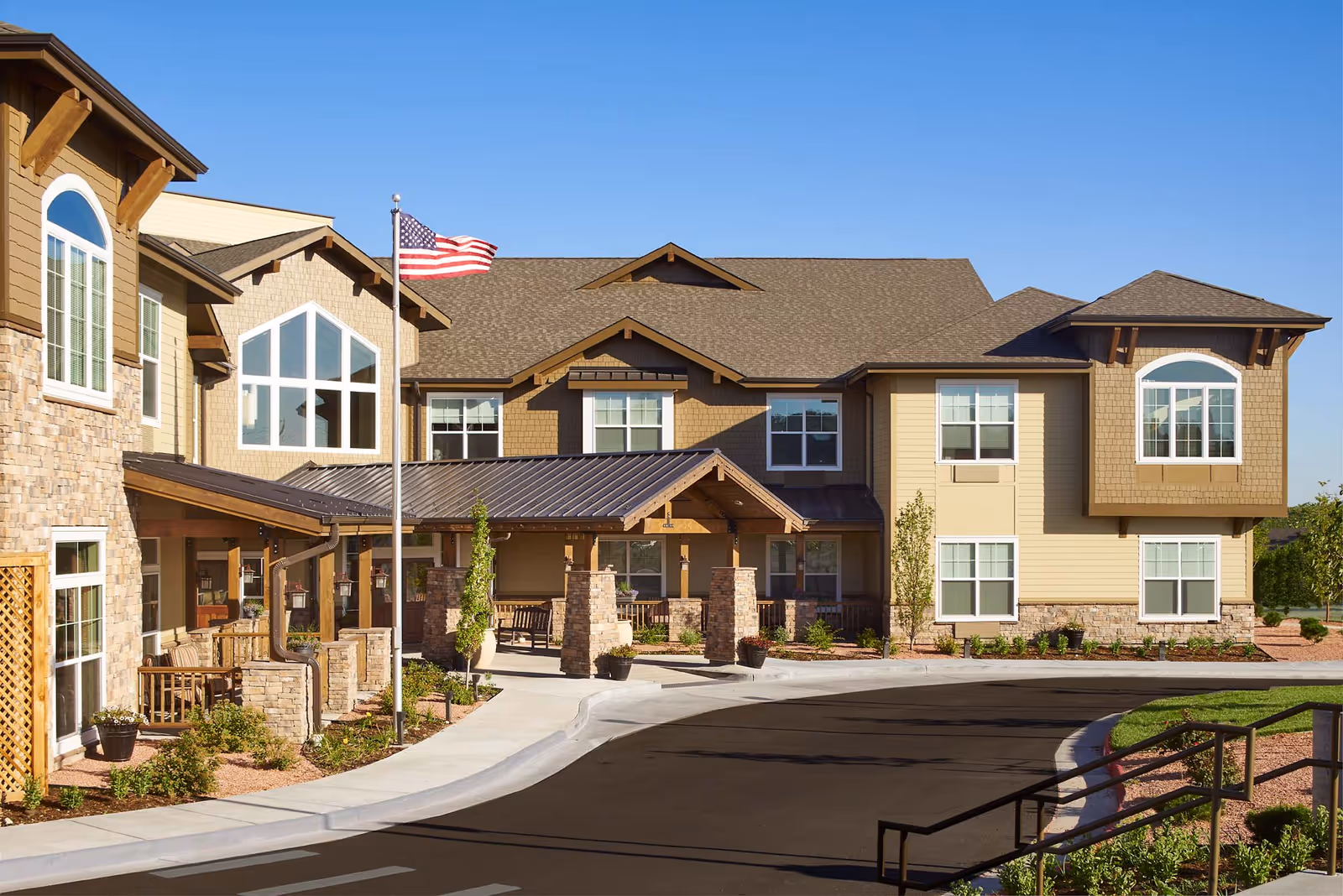 Exterior view of MorningStar Assisted Living & Memory Care of Wheat Ridge building with a clear blue sky, an American flag on a flagpole, stone and wood architectural details, and a paved driveway leading to the entrance.