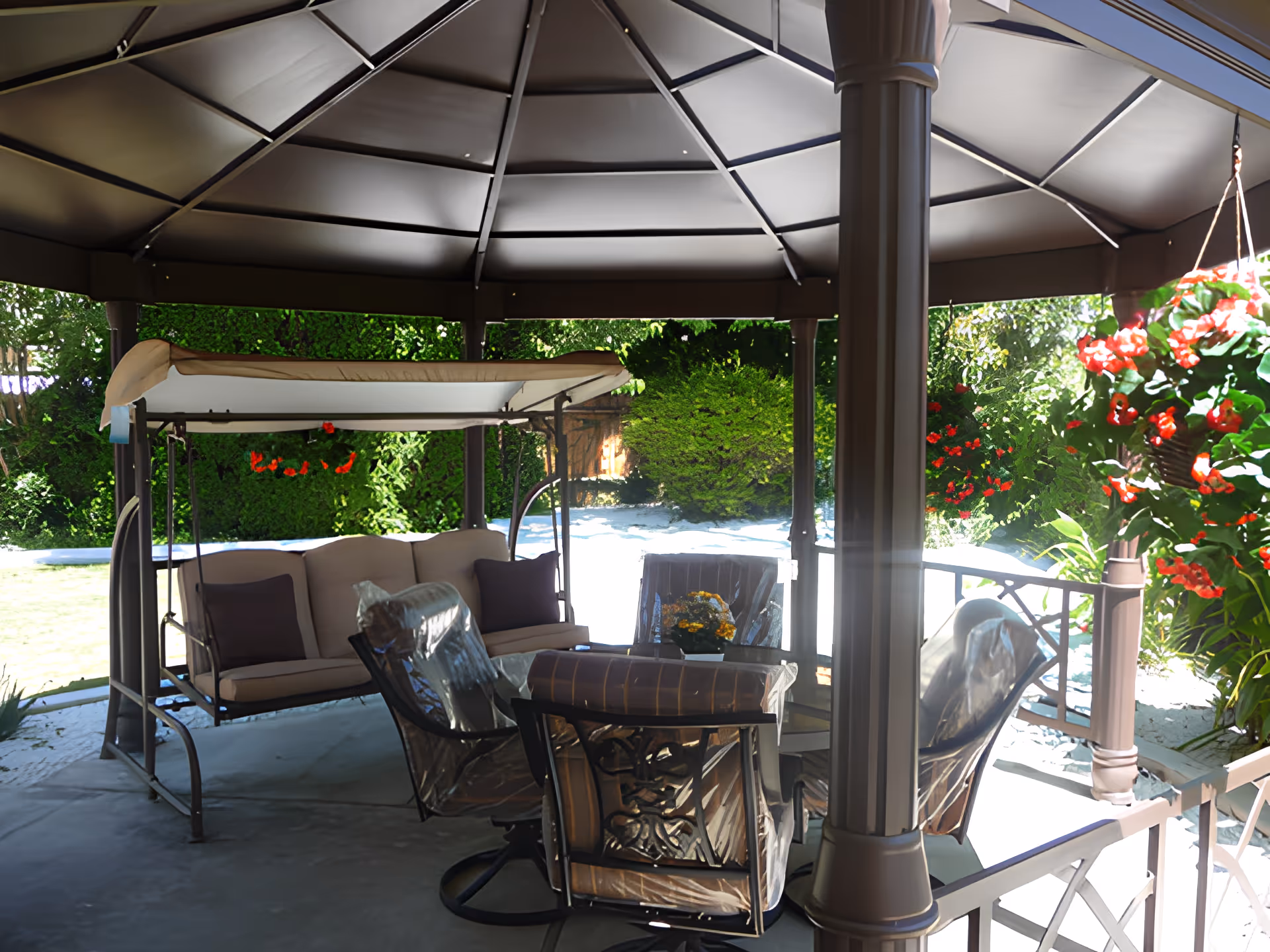 Covered gazebo patio with a cushioned swing, patio table and chairs, and hanging red flowers overlooking a backyard and pool.