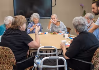 A group of elderly people sitting around a table in a communal room, engaging in conversation and eating snacks. A caregiver is standing nearby assisting them. A walker is visible near the table.