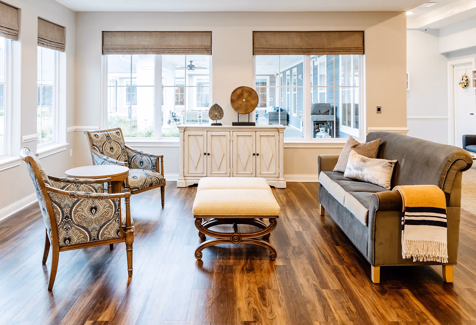 A bright and cozy living room area with two patterned armchairs and a small round table on the left, a cushioned ottoman in the center, and a dark gray sofa with pillows and a striped throw blanket on the right. Behind the furniture is a white cabinet with decorative items on top, and large windows with beige roman shades letting in natural light.