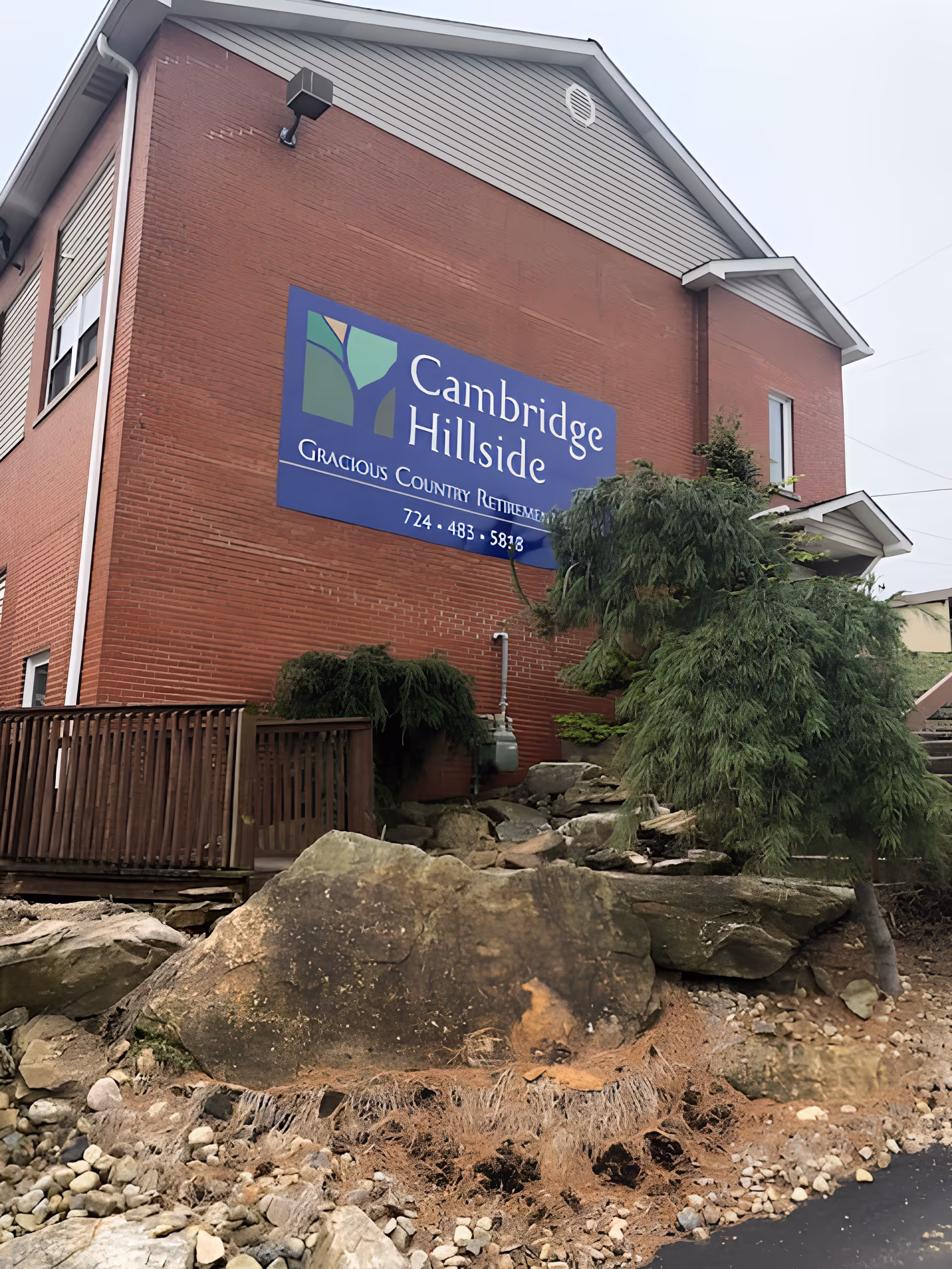 Exterior view of a red brick building with a large blue sign that reads 'Cambridge Hillside Gracious Country Retirement' along with a phone number. The building has a wooden ramp, some greenery, rocks, and a small tree in the foreground.