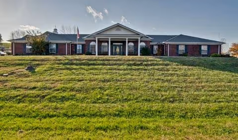 Front view of a single-story brick senior living building with a columned entrance and a large grassy lawn.