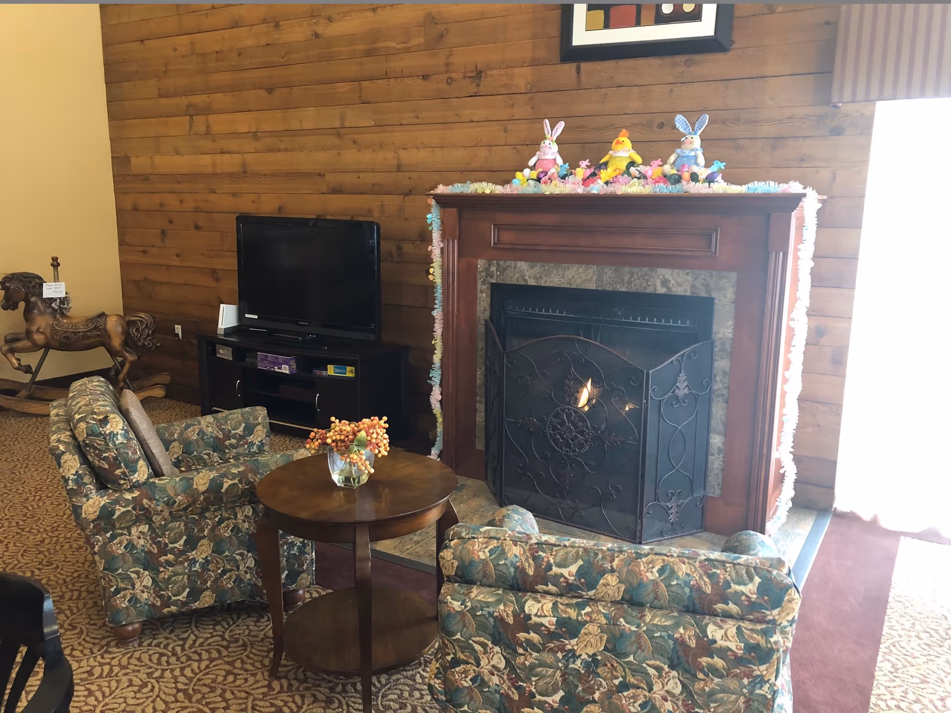A cozy living room area with two floral upholstered armchairs facing a wooden fireplace with a decorative metal screen. The fireplace mantle is decorated with colorful Easter-themed figurines including bunnies and chicks. To the left, there is a dark wooden TV stand with a flat-screen TV on top and a wooden rocking horse nearby. The walls are wooden paneled and the floor is carpeted with a patterned design.