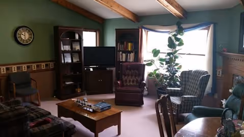 A cozy living room with exposed wooden ceiling beams, a large window with curtains, several armchairs including a burgundy leather chair and a plaid chair, a wooden coffee table with books and decorative items, a TV on a stand between two bookshelves, a clock on the green wall, and a fireplace on the right side.
