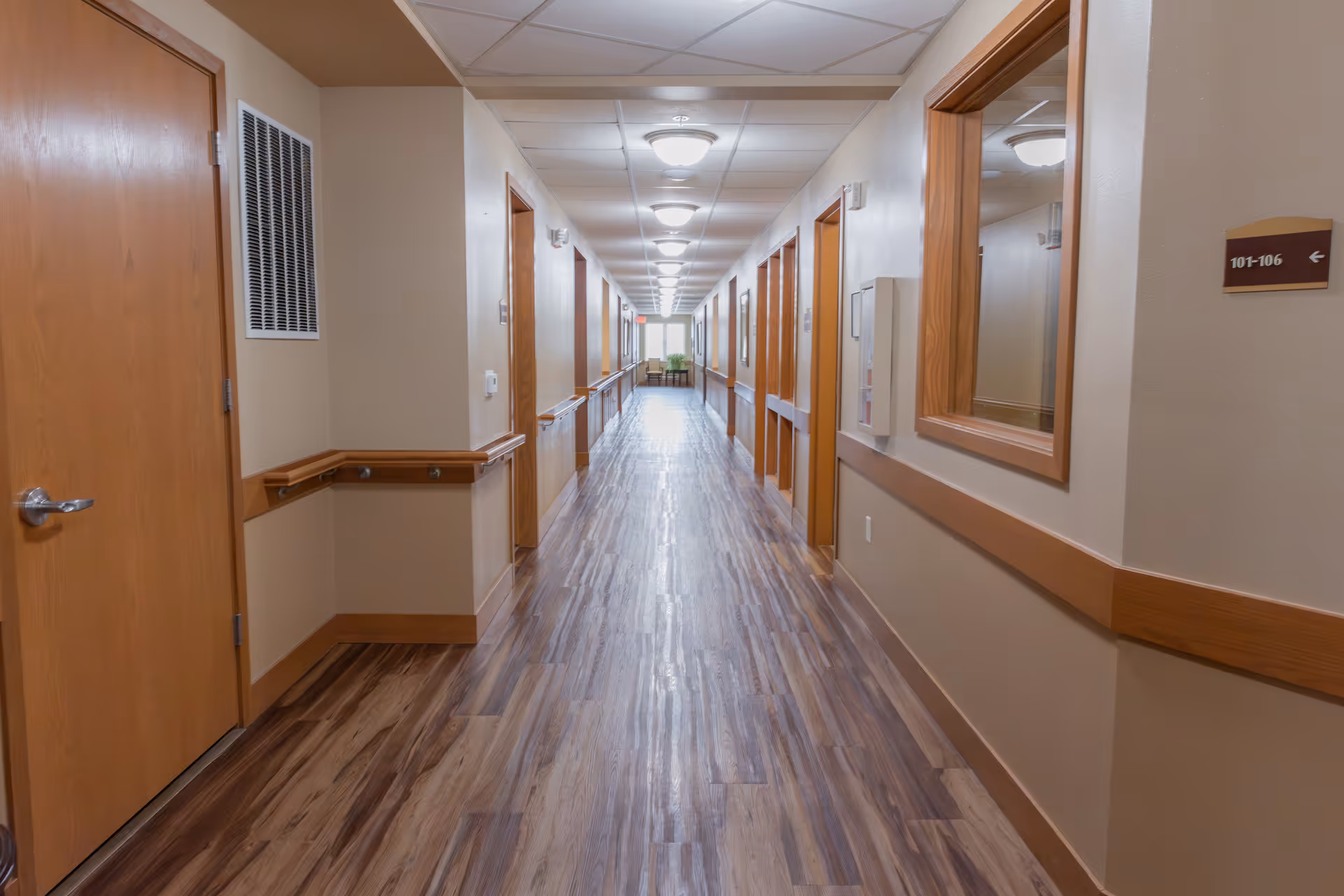 A long, well-lit hallway in a senior living facility with wood-patterned flooring, beige walls, wooden doors on both sides, handrails along the walls, ceiling lights, and a sign indicating rooms 101-106 to the right.