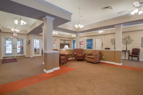 Interior view of a senior living facility common area with beige walls and carpet. There are several armchairs arranged near a half wall, a table lamp, and decorative wall art featuring tree designs. The area is well-lit with ceiling lights and has a door leading outside visible in the background.