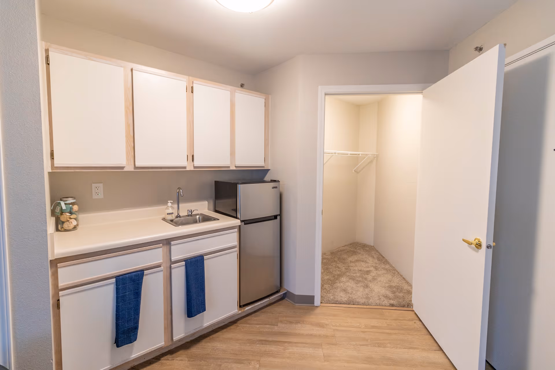 Small kitchenette area with white cabinets, a countertop with a sink, a small stainless steel refrigerator, and two blue towels hanging on the cabinet doors. To the right, there is an open door leading to a carpeted walk-in closet with a wire shelf.