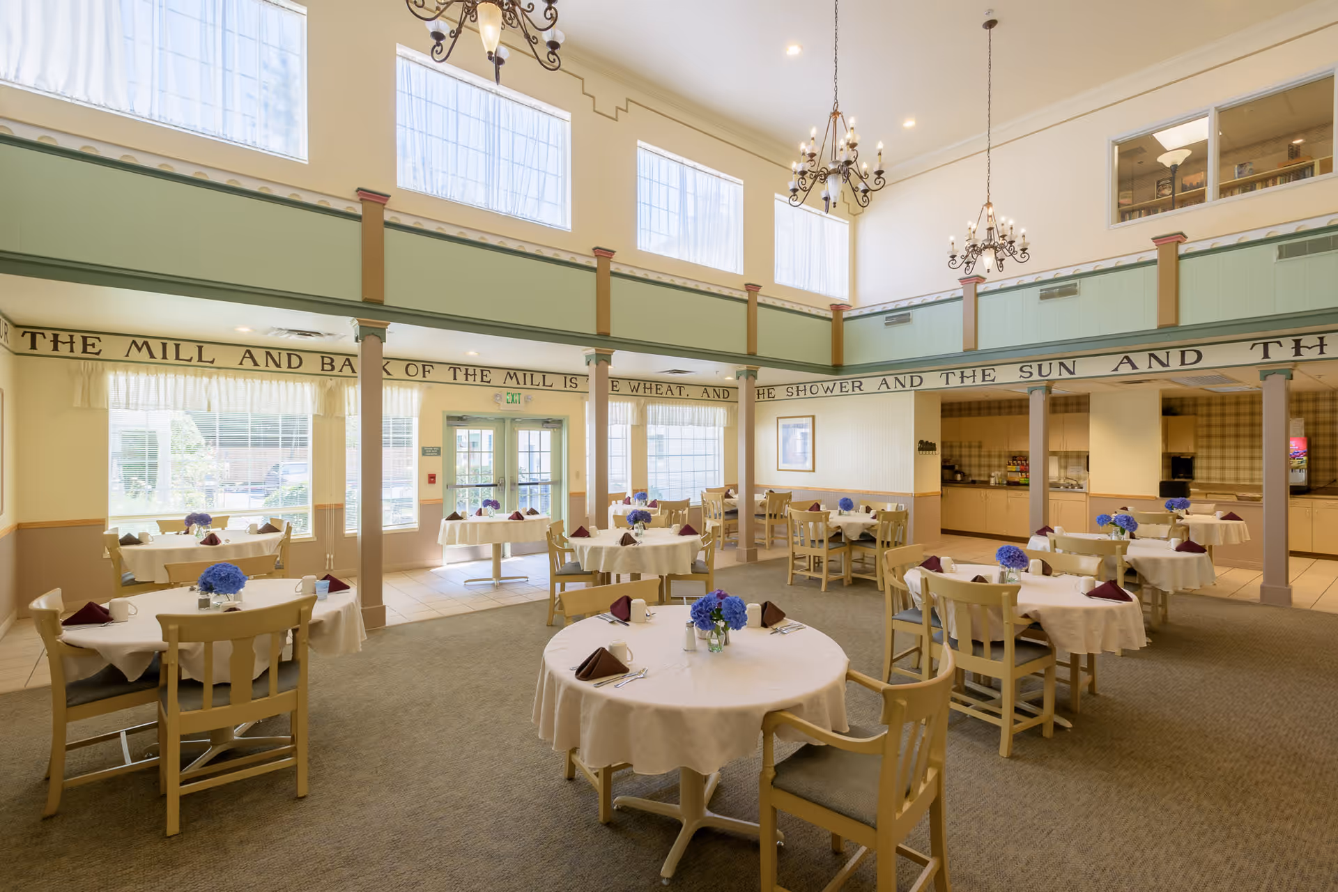 A bright and spacious dining room with multiple round tables covered in white tablecloths, each set with folded napkins and small floral centerpieces. The room features high ceilings with large windows near the top, allowing natural light to fill the space. Elegant chandeliers hang from the ceiling, and there is a serving area or kitchen visible in the background. The walls have a decorative trim with text running along the upper part of the room.