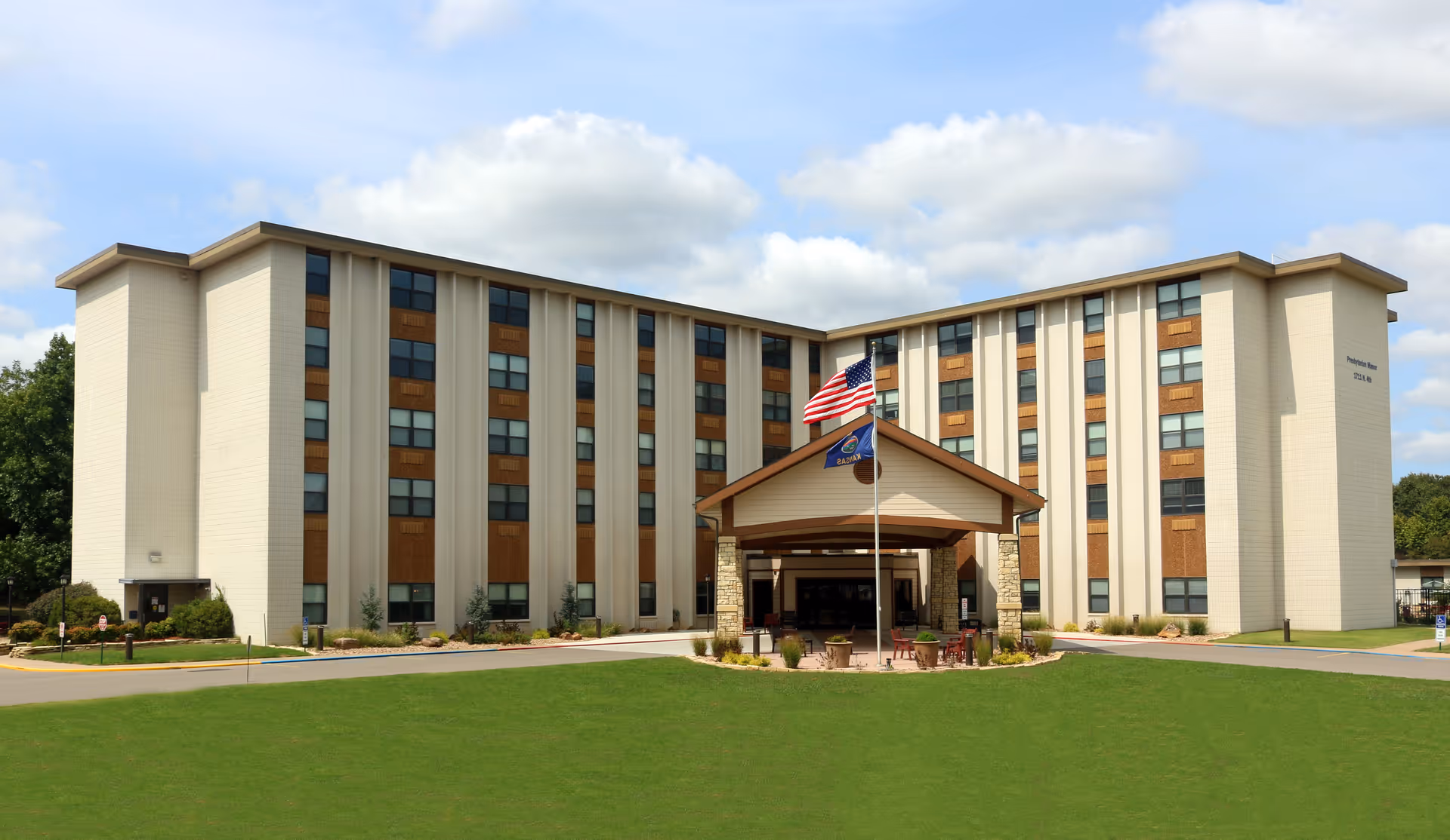Front view of Arkansas City Presbyterian Manor, a multi-story building with a covered entrance, flagpoles, and a large lawn.