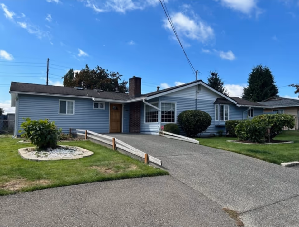 Single-story light blue house with a sloped roof, a chimney, and a wooden front door. The house has several windows and is surrounded by a well-maintained lawn with bushes and small garden beds. A concrete driveway with wooden railings leads up to the front entrance. The sky is clear with a few clouds.