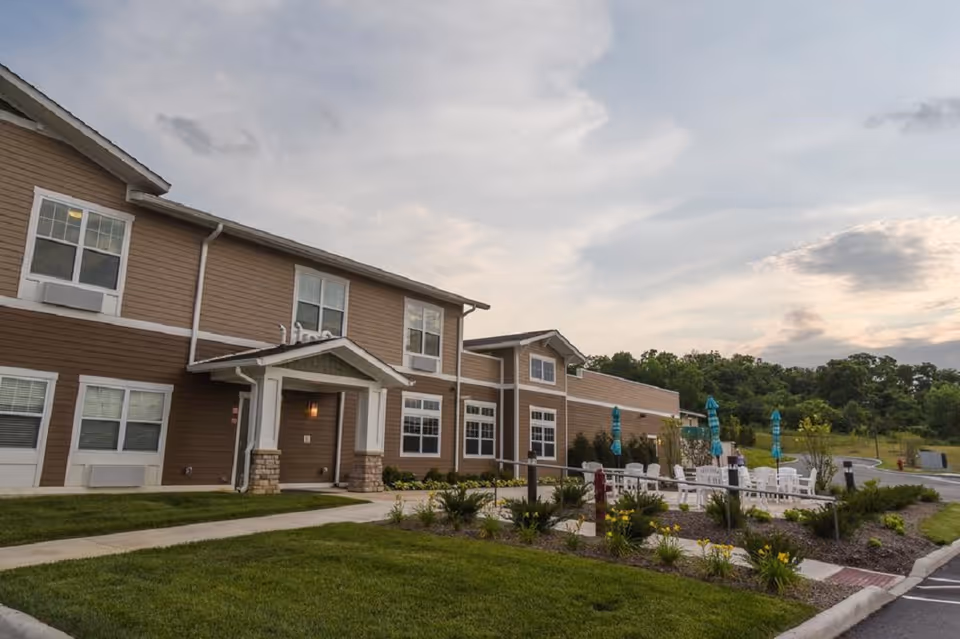 Two-story brown senior living building with a landscaped front lawn and an outdoor seating area with umbrellas under a cloudy sky.